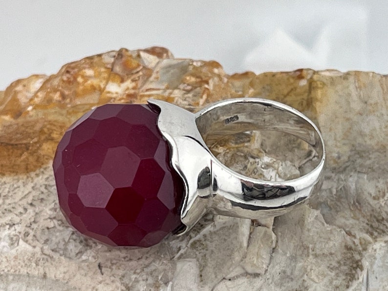 Op de afbeelding: Een zilveren ring met een grote, gefacetteerde, donkerrode edelsteen. De ring is in een eenvoudige, moderne stijl gezet.