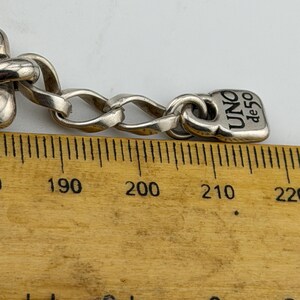 May include: Close-up of a silver chain bracelet with a rectangular tag that reads "UNO de 50". The chain links are oval-shaped. The bracelet is on a wooden ruler with centimetre markings.