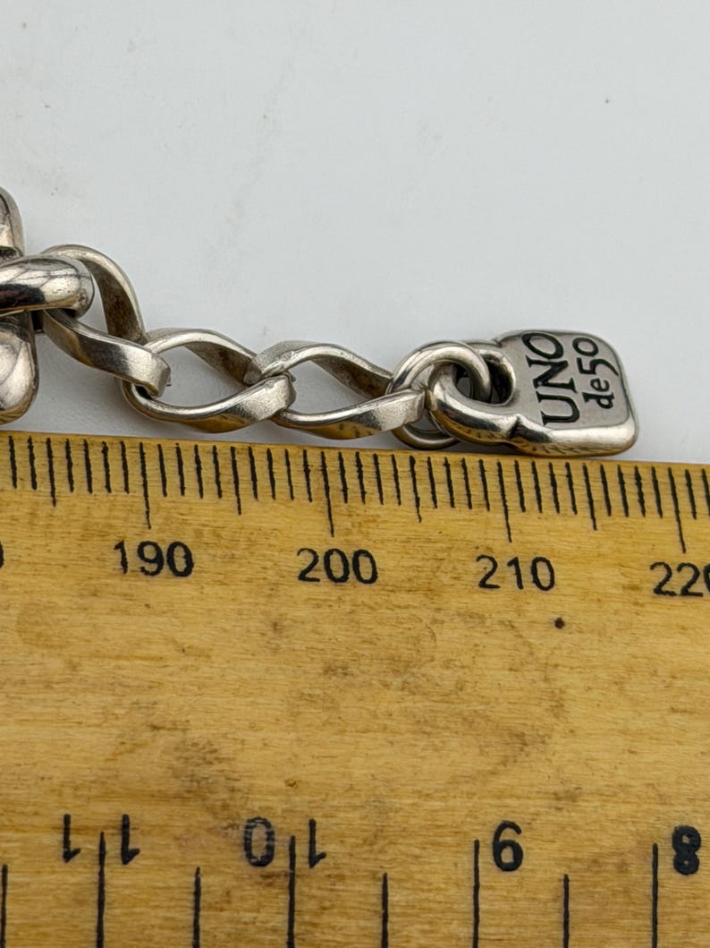 May include: Close-up of a silver chain bracelet with a rectangular tag that reads "UNO de 50". The chain links are oval-shaped. The bracelet is on a wooden ruler with centimetre markings.