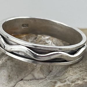 Pode incluir: Um anel de prata com um design de ondas. O anel tem um acabamento liso e polido e uma banda com um padrão ondulado ao redor do centro. O anel é provavelmente feito de prata esterlina, como indicado pelo selo "925".