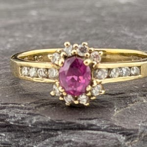 May include: A gold ring featuring a central oval-cut pink gemstone surrounded by a halo of small clear stones. The band includes a channel-set row of clear stones on each side, extending from the central cluster, creating a detailed design.