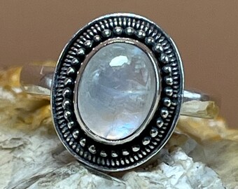 May include: A silver ring featuring a large oval moonstone set within a decorative bezel. The bezel is crafted from silver and has a beaded edge.
