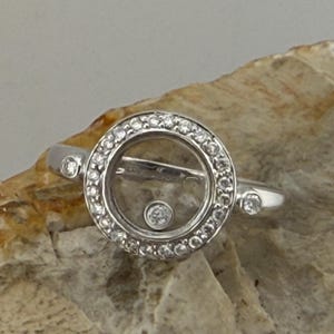 Puede incluir: Un anillo de plata con un diseño circular. El anillo presenta un centro transparente con un pequeño diamante flotante y está rodeado por un círculo de diamantes más pequeños. Se engarzan diamantes adicionales en la banda.