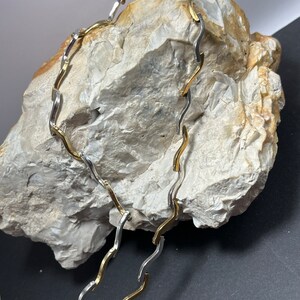 Op de afbeelding: Een gouden en zilveren ketting met een golvend ontwerp. De ketting ligt op een lichtgrijze steen.