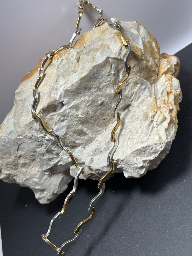 Op de afbeelding: Een gouden en zilveren ketting met een golvend ontwerp. De ketting ligt op een lichtgrijze steen.