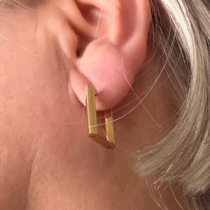 May include: A close-up of a gold-colored, rectangular earring. The earring is a simple, geometric design, with a rectangular shape. The earring is worn on the ear. The skin around the ear is visible.