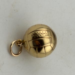 May include: A polished gold charm shaped like a volleyball, featuring a spherical design with embossed panel lines and a small circular jump ring attached to the side for hanging. The surface has a reflective metallic finish with clear detailing.