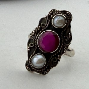 May include: Silver ring with an ornate design, featuring a large, round, magenta-colored gemstone in the center, flanked by two smaller, round, white pearls. The ring has a detailed, filigree-style setting.