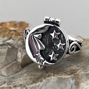 Peut inclure: Une bague en argent avec un médaillon rond à charnière représentant un croissant de lune et des étoiles. La bande de la bague est décorée d'un motif enroulé.