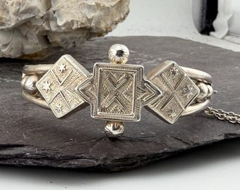 May include: A silver-toned metal cuff bracelet featuring a central square plaque with etched geometric patterns, flanked by two diamond-shaped panels decorated with star motifs and cross-hatching, accented by small spherical beads and a chain.