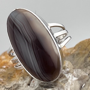 May include: A silver ring featuring a large oval agate stone. The stone has a dark brown base with white and grey stripes.
