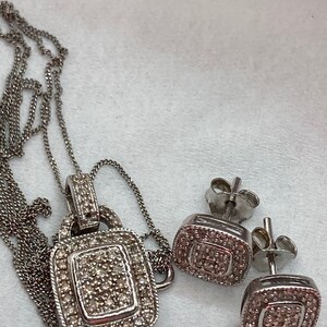 May include: A silver-toned jewelry set featuring a pendant necklace and matching stud earrings. The pendant and earrings have a square design, encrusted with small, sparkling stones. The necklace has a delicate chain.