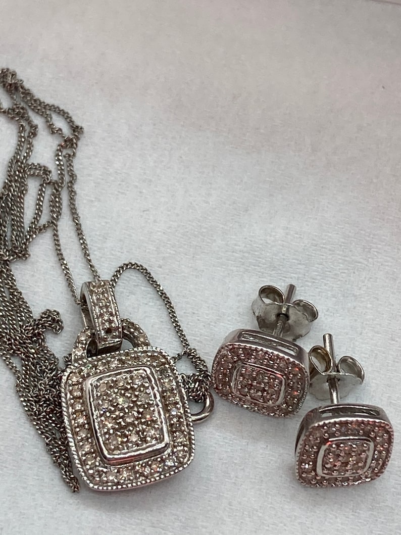 May include: A silver-toned jewelry set featuring a pendant necklace and matching stud earrings. The pendant and earrings have a square design, encrusted with small, sparkling stones. The necklace has a delicate chain.