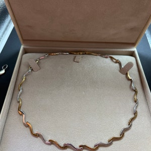 Op de afbeelding: Een gouden en zilveren ketting met een golvend ontwerp, gepresenteerd in een witte doos met de tekst "Chisholm Hunter Established 1863".
