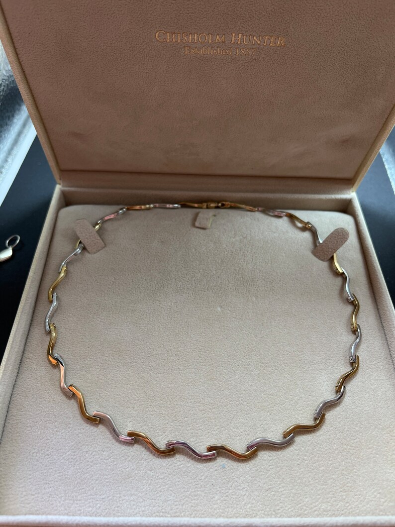 Op de afbeelding: Een gouden en zilveren ketting met een golvend ontwerp, gepresenteerd in een witte doos met de tekst "Chisholm Hunter Established 1863".