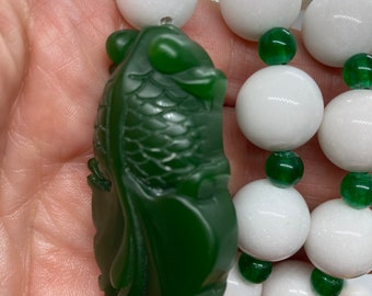 May include: A green jade fish pendant with intricate details hangs from a white and green beaded necklace. The necklace is made of round beads with a smooth, polished finish.