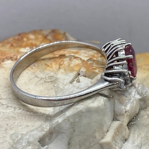 May include: A silver ring with a large, oval-shaped ruby gemstone surrounded by smaller diamonds. The ring is set on a white background.