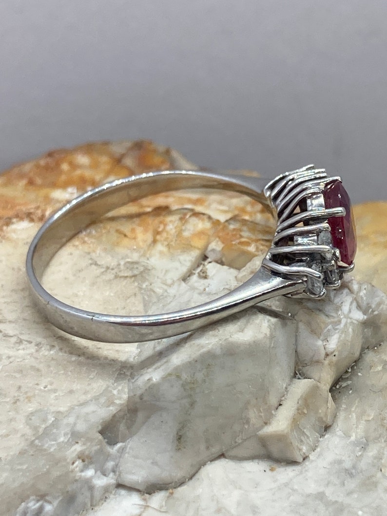 May include: A silver ring with a large, oval-shaped ruby gemstone surrounded by smaller diamonds. The ring is set on a white background.
