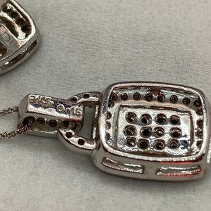 May include: Close-up of a silver pendant necklace with a square, gem-studded centerpiece. The pendant is attached to a delicate chain. The metal is stamped with "3KS" and "G015". The pendant is set with multiple small, dark gems.