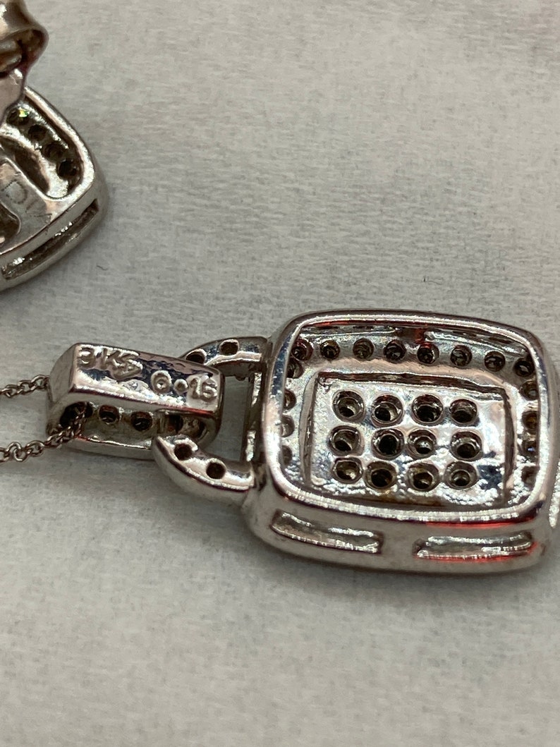 May include: Close-up of a silver pendant necklace with a square, gem-studded centerpiece. The pendant is attached to a delicate chain. The metal is stamped with "3KS" and "G015". The pendant is set with multiple small, dark gems.