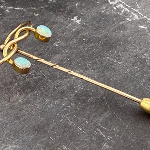 Op de afbeelding: Een goudkleurige stropdaspen met twee ovale, opalescerende stenen en een decoratieve bovenkant. De speld heeft een lange, slanke schacht met een spiraalvormig ontwerp en een afgerond uiteinde. De opalescerende stenen hebben een lichtblauwe en witte kleur.