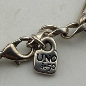 May include: Close-up of a silver-toned necklace clasp and chain. The clasp is a lobster claw style, and the chain links are oval. A small, rectangular charm with the text "UNO de 50" is attached to the chain.