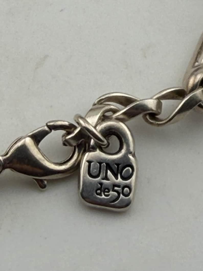 May include: Close-up of a silver-toned necklace clasp and chain. The clasp is a lobster claw style, and the chain links are oval. A small, rectangular charm with the text "UNO de 50" is attached to the chain.