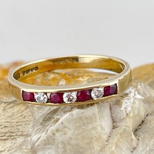 Op de afbeelding: Een gouden bandring met afwisselend rode en heldere edelstenen. De ring is bezet met vijf rode robijnen en vier heldere diamanten. De ring wordt gepresenteerd op een getextureerd, lichtgekleurd oppervlak.