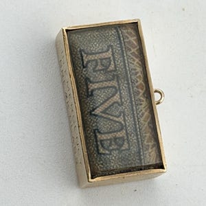 Puede incluir: Colgante rectangular dorado con una cara cubierta de vidrio. La cara muestra la palabra "FIVE" en letras oscuras sobre un fondo estampado. El colgante tiene un pequeño lazo para una cadena.