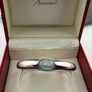 May include: A silver-toned bracelet with a light blue, oval-shaped center stone, presented in a red and white box. The box lid is open, revealing the bracelet resting on a white cushion. The word "Baccarat" is printed on the inside of the box lid.