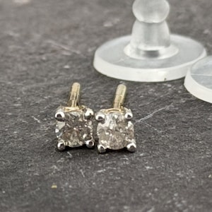 May include: A pair of square-cut diamond stud earrings with gold posts and settings. The diamonds are set in a four-prong setting. The earrings are displayed on a dark gray surface with two clear earring backs.