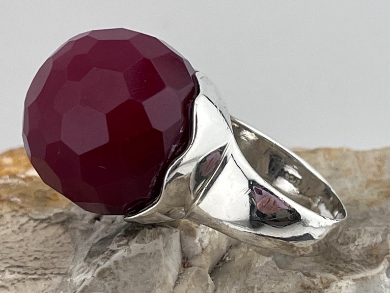 Op de afbeelding: Een zilveren ring met een grote, gefacetteerde, bordeauxrode edelsteen. De ring heeft een uniek, golvend ontwerp.