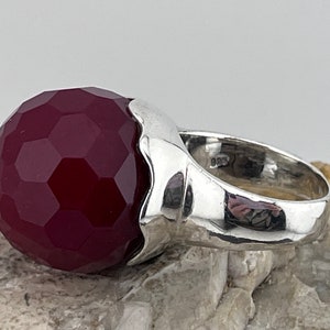 Op de afbeelding: Een zilveren ring met een grote, gefacetteerde, donkerrode edelsteen. De ring heeft een getextureerde band.