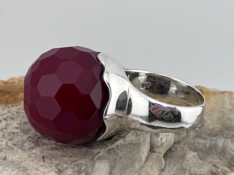 Op de afbeelding: Een zilveren ring met een grote, gefacetteerde, donkerrode edelsteen. De ring heeft een getextureerde band.