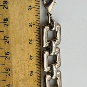 May include: Silver bracelet with rectangular links, next to a wooden ruler. The bracelet's links are approximately 1.5 cm wide. The clasp is a lobster claw style.