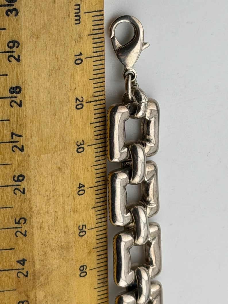 May include: Silver bracelet with rectangular links, next to a wooden ruler. The bracelet's links are approximately 1.5 cm wide. The clasp is a lobster claw style.