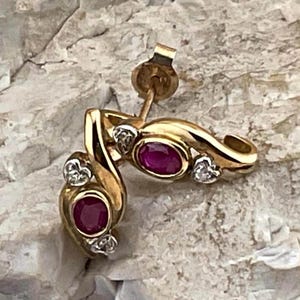 May include: A pair of gold stud earrings featuring a curved, polished metal design. Each earring is set with an oval-cut deep red ruby and two small, clear, round-cut accent stones. The earrings include a standard post and butterfly back closure.