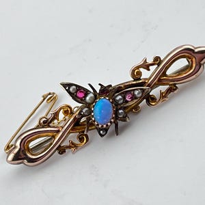 Peut inclure: Une broche en or représentant un papillon stylisé avec une pierre centrale en opale bleue et des accents de pierres roses et blanches. La broche est montée sur un motif délicat et tourbillonnant.