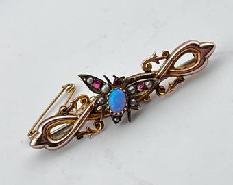 May include: A gold brooch featuring a stylized butterfly with a blue opal center stone and accents of pink and white stones. The brooch is set on a delicate, swirling design.