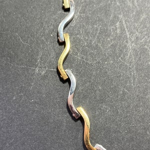 Op de afbeelding: Een close-up van een gouden en zilveren ketting met een golvend ontwerp. De ketting is gemaakt van in elkaar grijpende schakels die afwisselend goud en zilver zijn.