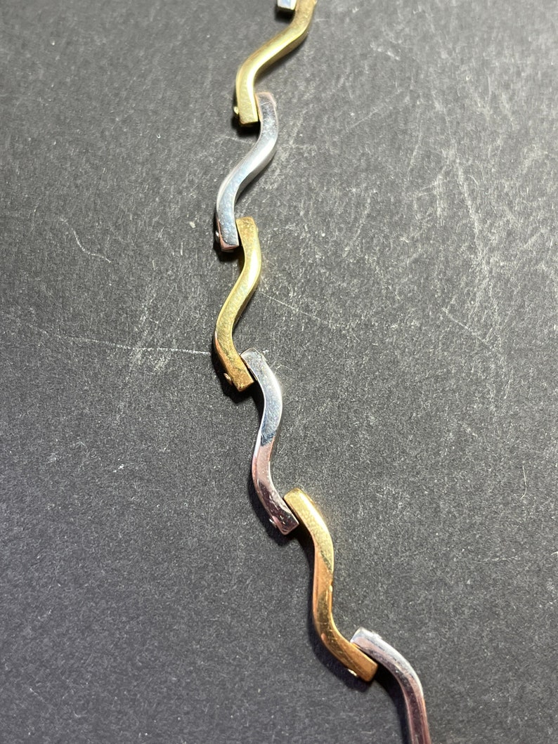 Op de afbeelding: Een close-up van een gouden en zilveren ketting met een golvend ontwerp. De ketting is gemaakt van in elkaar grijpende schakels die afwisselend goud en zilver zijn.
