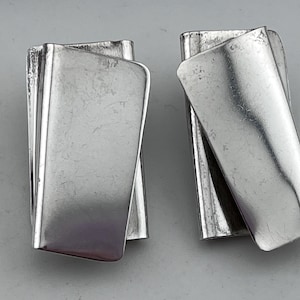 Op de afbeelding: Twee zilverkleurige metalen oorbellen. Elke oorbel heeft een rechthoekige vorm met een gebogen rand en een gevouwen ontwerp. De oorbellen hebben een geborstelde metalen afwerking tegen een grijze achtergrond.