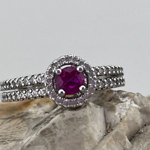 May include: A silver ring featuring a round, deep pink ruby set within a halo of smaller diamonds. The ring has a double band of smaller diamonds.