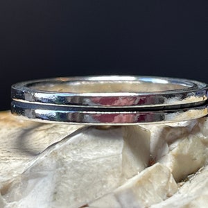 Puede incluir: Un anillo de plata con un diseño de doble banda. El anillo tiene una superficie pulida y reflectante y una ranura oscura entre las dos bandas. El anillo se muestra sobre una superficie clara y texturizada sobre un fondo oscuro.