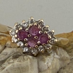 Può includere: Un anello d'oro con un gruppo di pietre preziose rosa circondate da piccole pietre bianche. L'anello ha un design floreale, con le pietre rosa al centro e le pietre bianche che formano un alone. Sfondo neutro.