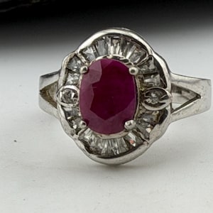 Könnte beinhalten: Ein silberfarbener Ring mit einem oval geschliffenen, dunkelroten Edelstein, der von vier Krappen gehalten wird. Der Mittelstein ist von einem Kranz aus klaren Baguette-Steinen und zwei kleinen runden, klaren Steinen an den Seiten umgeben.