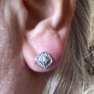 May include: A silver stud earring with a square, textured design. The earring is encrusted with small, sparkling stones, creating a shimmering effect. The earring is set against the backdrop of a person's ear and blonde hair.