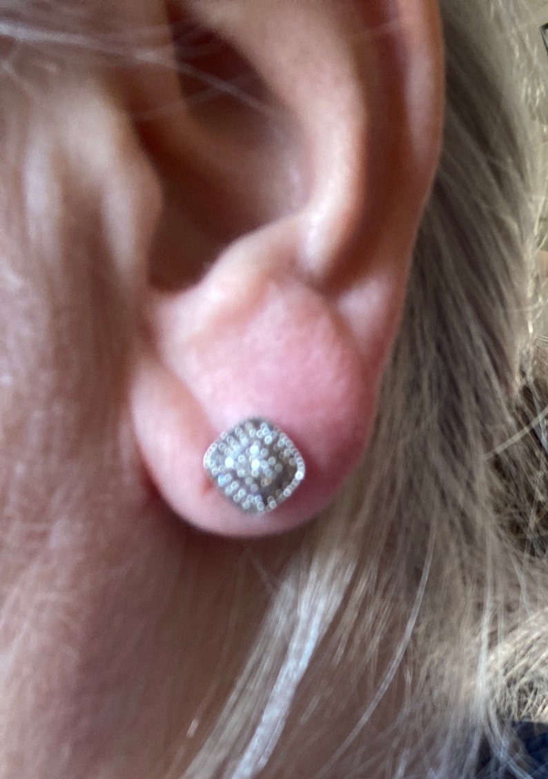 May include: A silver stud earring with a square, textured design. The earring is encrusted with small, sparkling stones, creating a shimmering effect. The earring is set against the backdrop of a person's ear and blonde hair.