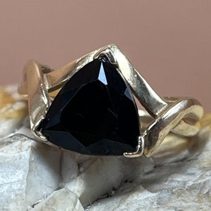 Könnte beinhalten: Ein silberner Ring mit einem schwarzen dreieckigen Edelstein, der in der Mitte eingesetzt ist. Der Ring hat ein einzigartiges, verdrehtes Design.