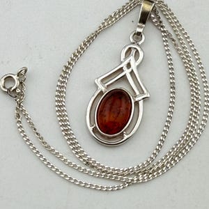 May include: A silver pendant necklace featuring an oval-shaped, deep orange amber gemstone set within a polished silver frame. The pendant hangs from a delicate silver chain with a circular clasp, showcasing a clean, metallic finish and warm stone.
